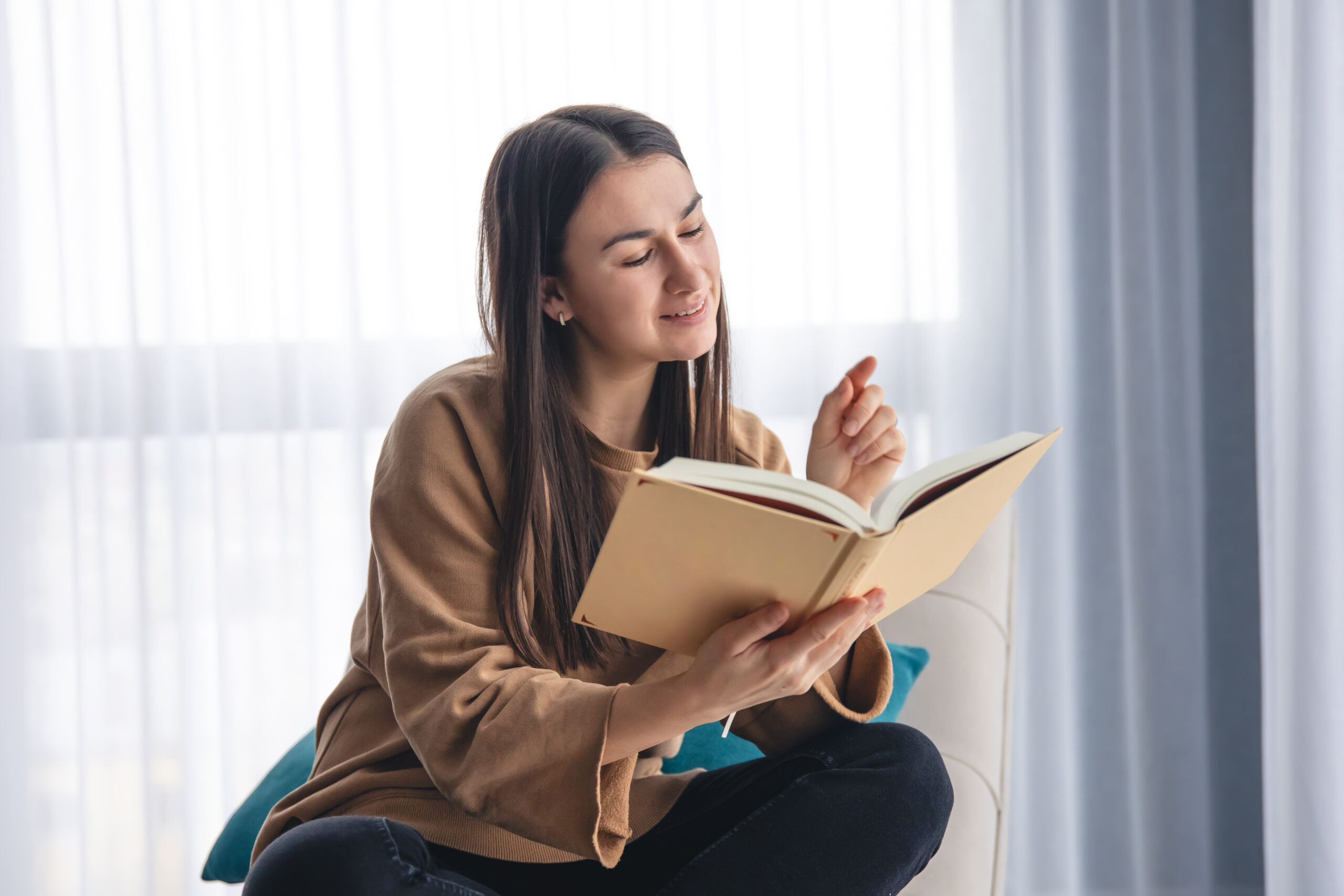 A young woman is reading a book while sitting on an armchair by the window.